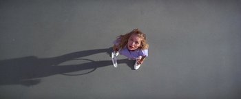 Movie still from “The Witches of Eastwick” (1987), directed by George Miller – A woman holding two tennis rackets on a tennis court; Wide shot, Overhead angle