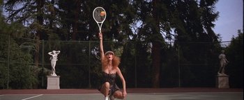 Movie still from “The Witches of Eastwick” (1987), directed by George Miller – A female tennis player in a polka dot dress is holding a tennis ball and swinging a tennis racket; Wide shot, Low angle