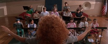 Movie still from “The Witches of Eastwick” (1987), directed by George Miller – A group of people sitting in front of an audience; Wide shot, Over the shoulder angle
