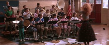 Movie still from “The Witches of Eastwick” (1987), directed by George Miller – A group of people sitting in a room playing musical instruments; Wide shot, High angle