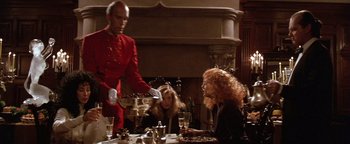 Movie still from “The Witches of Eastwick” (1987), directed by George Miller – A man in a red uniform serving a woman at a dinner table; Medium shot, Low angle