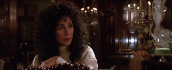Movie still from “The Witches of Eastwick” (1987), directed by George Miller – A person with curly hair near a table; Close Up shot, Over the shoulder angle