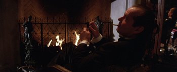 Movie still from “The Witches of Eastwick” (1987), directed by George Miller – A man sitting in front of a fireplace smoking a cigarette; Medium shot, Low angle