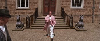 Movie still from “The Witches of Eastwick” (1987), directed by George Miller – A man in a pink coat walking in front of a building; Wide shot, Low angle