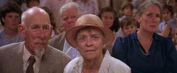 Movie still from “The Witches of Eastwick” (1987), directed by George Miller – An older woman wearing a tan hat in front of a group of people; Close Up shot, Low angle