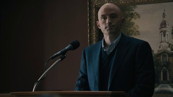 Movie still from “The Wizard of Lies” (2017), directed by Barry Levinson – A bald man in a suit is standing at a podium; Medium shot, Over the shoulder angle