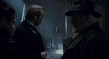 Movie still from “The Wolfman” (2010), directed by Joe Johnston – Two men in suits and hats talking in a dimly - lit room; Medium shot, Over the shoulder angle