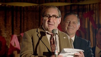 Movie still from “The World's Fastest Indian” (2005), directed by Roger Donaldson – A man in a brown suit and glasses speaking at a microphone; Close Up shot, Over the shoulder angle