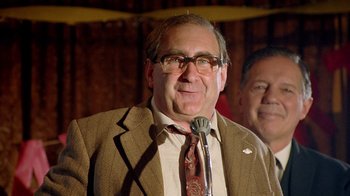 Movie still from “The World's Fastest Indian” (2005), directed by Roger Donaldson – An older man wearing glasses and a suit and tie while speaking into a microphone; Close Up shot, Low angle