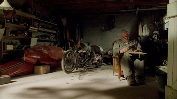 Movie still from “The World's Fastest Indian” (2005), directed by Roger Donaldson – A man sitting in a chair in front of a motorcycle in a garage; Wide shot, High angle