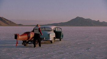 Movie still from “The World's Fastest Indian” (2005), directed by Roger Donaldson – A man with a surfboard next to an old car; Extreme Wide shot, High angle