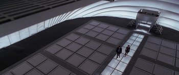 Movie still from “The X Files” (1998), directed by Rob Bowman – Two people are standing on a platform in the middle of a building; Extreme Wide shot, Overhead angle