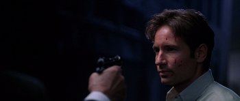 Movie still from “The X Files” (1998), directed by Rob Bowman – A man holding a gun in his hand and looking at the camera; Close Up shot, Over the shoulder angle