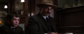 Movie still from “There Will Be Blood” (2007), directed by Paul Thomas Anderson – A man in a suit and a hat is sitting at a table; Close Up shot, Low angle