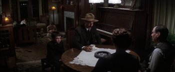 Movie still from “There Will Be Blood” (2007), directed by Paul Thomas Anderson – A man in a suit and hat sitting at a table with two other people; Medium shot, Over the shoulder angle