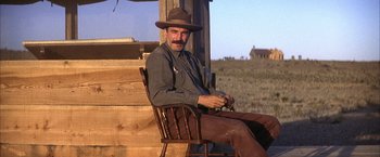 Movie still from “There Will Be Blood” (2007), directed by Paul Thomas Anderson – A man sitting on a wooden chair in front of a building; Medium shot, Low angle