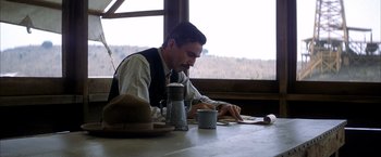Movie still from “There Will Be Blood” (2007), directed by Paul Thomas Anderson – A man sitting at a table writing on a piece of paper; Medium shot, High angle
