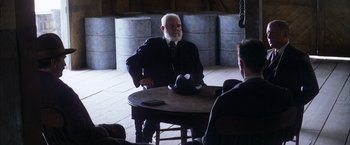Movie still from “There Will Be Blood” (2007), directed by Paul Thomas Anderson – An old man sitting at a round table wearing a hat; Medium shot, Over the shoulder angle