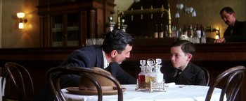 Movie still from “There Will Be Blood” (2007), directed by Paul Thomas Anderson – A man sitting at a table in a restaurant with another man; Medium shot, Over the shoulder angle