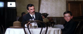 Movie still from “There Will Be Blood” (2007), directed by Paul Thomas Anderson – A man sitting at a table with a glass of wine; Medium shot, Over the shoulder angle
