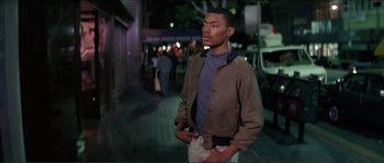 Movie still from “They Live” (1988), directed by John Carpenter – A man standing on the sidewalk at night with his hands in his pockets; Medium shot, Over the shoulder angle