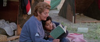Movie still from “They Live” (1988), directed by John Carpenter – An older woman sitting next to a young boy reading a book; Medium shot, Over the shoulder angle