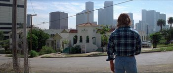 Movie still from “They Live” (1988), directed by John Carpenter – A man standing on the side of the road in front of a building; Wide shot, Over the shoulder angle