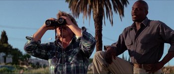 Movie still from “They Live” (1988), directed by John Carpenter – A man looking through binoculars while standing next to another man; Medium shot, Low angle