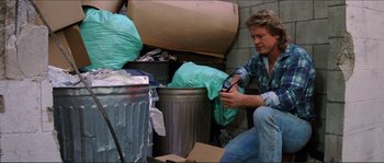 Movie still from “They Live” (1988), directed by John Carpenter – A man sitting on the ground next to a trash can; Medium shot, Low angle