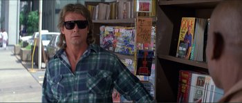 Movie still from “They Live” (1988), directed by John Carpenter – A man wearing a plaid shirt and sunglasses in front of a book case; Medium shot, Low angle