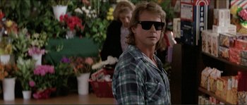 Movie still from “They Live” (1988), directed by John Carpenter – A man wearing sunglasses standing next to flowers; Medium shot, Over the shoulder angle