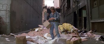 Movie still from “They Live” (1988), directed by John Carpenter – A man standing on a pile of cardboard looking at a box; Wide shot, Low angle