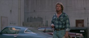 Movie still from “They Live” (1988), directed by John Carpenter – A man in plaid shirt holding a cell phone in a parking lot; Medium shot, Low angle