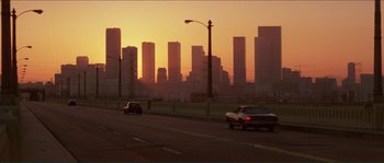 Movie still from “They Live” (1988), directed by John Carpenter – Cars driving down a street in front of a city skyline at sunset; Extreme Wide shot, High angle