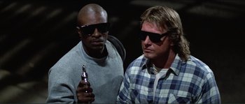 Movie still from “They Live” (1988), directed by John Carpenter – Two men with sunglasses are holding a cell phone; Medium shot, Low angle