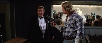 Movie still from “They Live” (1988), directed by John Carpenter – A man holding a gun in his hand while another man stands next to him in front of a window; Medium shot, Over the shoulder angle