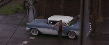 Movie still from “This Boy's Life” (1993), directed by Michael Caton-Jones – A man standing next to an old car holding a bouquet of flowers; Wide shot, High angle
