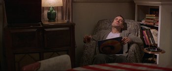 Movie still from “This Boy's Life” (1993), directed by Michael Caton-Jones – A man sitting in a chair holding a book; Medium shot, High angle