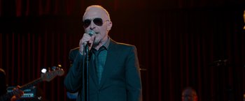 Movie still from “This Is 40” (2012), directed by Judd Apatow – An older man in a suit and sunglasses is holding a microphone; Close Up shot, Low angle