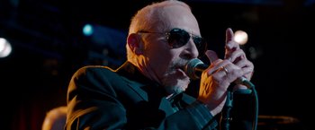 Movie still from “This Is 40” (2012), directed by Judd Apatow – An older man wearing sunglasses and a black jacket is holding a microphone; Close Up shot, Low angle