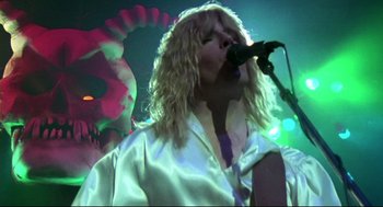 Movie still from “This Is Spinal Tap” (1984), directed by Rob Reiner – A person with long blonde curly hair is holding a microphone; Close Up shot, Low angle