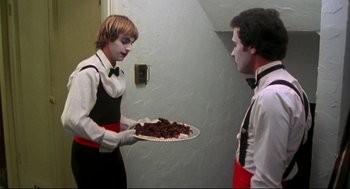 Movie still from “This Is Spinal Tap” (1984), directed by Rob Reiner – A man holding a plate of food next to another man; Medium shot, Over the shoulder angle