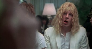Movie still from “This Is Spinal Tap” (1984), directed by Rob Reiner – A man with long blonde hair and a white suit jacket; Close Up shot, Over the shoulder angle