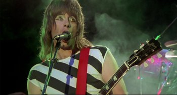 Movie still from “This Is Spinal Tap” (1984), directed by Rob Reiner – A man in a striped shirt holding a microphone and a guitar; Close Up shot, Low angle