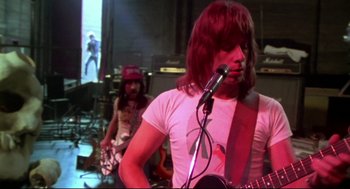 Movie still from “This Is Spinal Tap” (1984), directed by Rob Reiner – A man with long brown hair is holding a microphone; Medium shot, Low angle