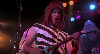 Movie still from “This Is Spinal Tap” (1984), directed by Rob Reiner – A man in striped shirt playing guitar on stage; Medium shot, Low angle