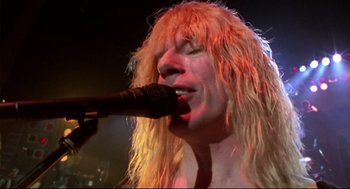 Movie still from “This Is Spinal Tap” (1984), directed by Rob Reiner – A man with long blonde hair is holding a microphone up to his mouth; Close Up shot, Low angle
