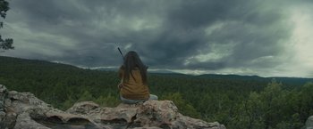 Movie still from “Those Who Wish Me Dead” (2021), directed by Taylor Sheridan – A woman sitting on top of a mountain holding a stick; Wide shot, Low angle