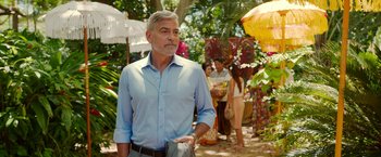 Movie still from “Ticket to Paradise” (2022), directed by Ol Parker – A man in a blue dress shirt is standing in the grass; Medium shot, Over the shoulder angle