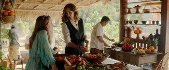 Movie still from “Ticket to Paradise” (2022), directed by Ol Parker – A group of people standing around a table with food on top of it; Medium shot, Over the shoulder angle